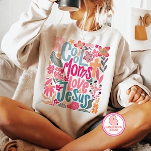 May include: White sweatshirt with a colorful graphic design that says "Cool Moms Love Jesus". The design features flowers, leaves, and a cross.
