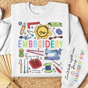 Puede incluir: Una sudadera gris con un diseño de bordado colorido que incluye una máquina de coser, agujas, hilo y el texto "EMBROIDERY". El texto "Can't talk right now Doing EMBROIDERY STUFF" está en la manga derecha.