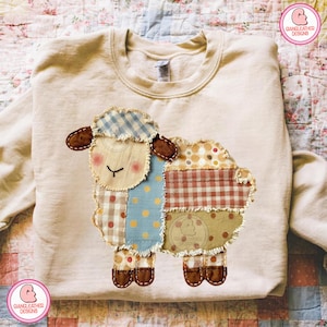 Peut inclure: Un sweat-shirt beige orné d'un mouton en patchwork. Le mouton porte un chapeau à carreaux bleu et blanc, des oreilles marron et un visage aux joues roses. Le corps est fait de divers tissus à motifs, dont des pois et du vichy.