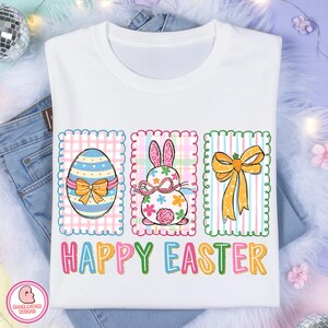 May include: White t-shirt with a colorful Easter design. The design includes an Easter egg, a bunny, and a bow, each within a decorative frame. The words "HAPPY EASTER" are printed in colorful letters below the images.