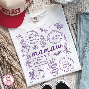 May include: White t-shirt with purple floral illustrations and the word "mamaw". The design includes phrases like "spoil them sweet" and "cherish every moment". A red and white baseball cap is visible.