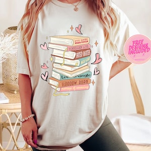 May include: A white t-shirt with a stack of books printed on the front. The books have different titles, including "Soul Mates", "Forbidden Love", "Fated Mates", "Age Gap", "Love Triangle", "Slooooow Burn", and "Marriage of Convenience".