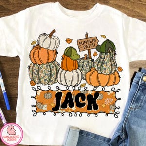May include: White t-shirt featuring a colorful pumpkin patch design. The design includes stacked pumpkins in orange, white, and camouflage patterns, with a sign that reads "Pumpkin Patch" and the name "Jack".