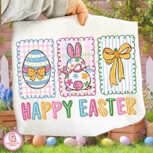 May include: A white Easter-themed towel featuring three colorful panels. The panels display an Easter egg, a bunny, and a bow, all with decorative borders. The words "HAPPY EASTER" are printed across the bottom in bright colors.