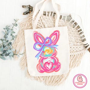 May include: A white tote bag with a colorful abstract bunny design. The bunny is pink, blue, yellow, and green. The bunny has a large blue bow on its head.