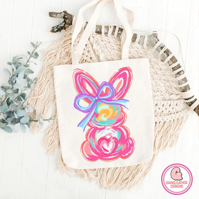 May include: A white tote bag with a colorful abstract bunny design. The bunny is pink, blue, yellow, and green. The bunny has a large blue bow on its head.