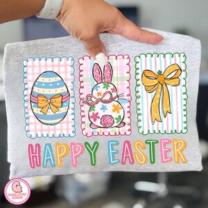 May include: A light gray t-shirt featuring three Easter-themed designs. The designs include a decorated egg, a bunny with floral accents, and a carrot bow. The words "HAPPY EASTER" are printed in colorful letters below the designs.