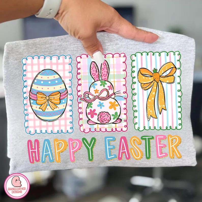 May include: A light gray t-shirt featuring three Easter-themed designs. The designs include a decorated egg, a bunny with floral accents, and a carrot bow. The words "HAPPY EASTER" are printed in colorful letters below the designs.