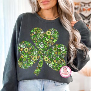 May include: Dark gray sweatshirt featuring a large shamrock design filled with green, white, and yellow floral patterns. The shamrock has a heart-shaped top. The sweatshirt has long sleeves and a crew neck. The design includes the text "GIANGLEATHER DESIGNS".