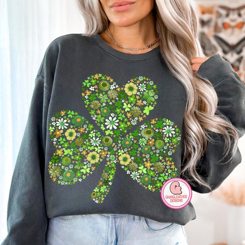 May include: Dark gray sweatshirt featuring a large shamrock design filled with green, white, and yellow floral patterns. The shamrock has a heart-shaped top. The sweatshirt has long sleeves and a crew neck. The design includes the text "GIANGLEATHER DESIGNS".