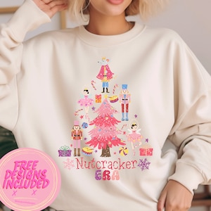 May include: A white sweatshirt with a pink and white Christmas tree design featuring a Nutcracker theme. The design includes a Nutcracker soldier, a mouse king, and ballerinas. The text "Nutcracker Era" is printed below the tree.