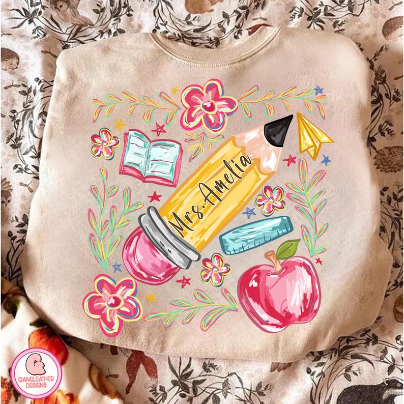 May include: Beige sweatshirt with a colorful teacher-themed design. A large yellow pencil with "Mrs. Amelia" printed on it is surrounded by flowers, a red apple, an open book, and a ruler. A paper airplane and stars complete the design.
