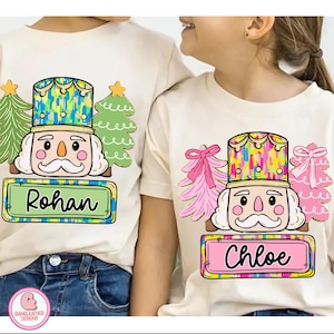 May include: Two cream-colored t-shirts with a colorful nutcracker design and the names "Rohan" and "Chloe" on rectangular nameplates. The design includes Christmas trees and a decorative hat. The shirts are likely for children.
