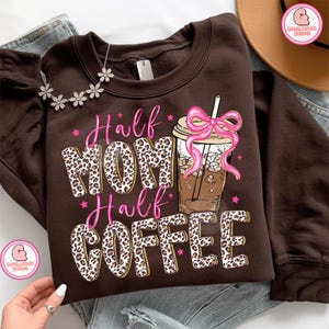 May include: A brown sweatshirt with the text "Half MOM Half COFFEE" in leopard print and pink lettering. A graphic of an iced coffee with a pink bow is on the right. A floral necklace is draped on the shoulder.