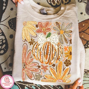 May include: A beige sweatshirt with a vibrant autumn design. The print showcases a pumpkin with a bow, encircled by orange, gold, and brown flowers. The design incorporates a glitter effect for added detail.