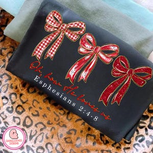 May include: Black t-shirt featuring three red bows with gold trim. The bows have different patterns, including gingham and glitter. The text "Oh how He loves us Ephesians 2:4-8" is written in red script. The shirt is folded.