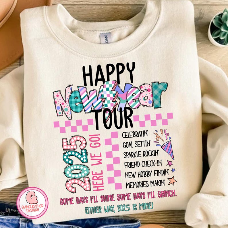 May include: A cream-colored sweatshirt with a colorful graphic design that says "Happy New Year Tour 2025 Here We Go!" The graphic includes a pink and white checkered pattern, a colorful "Happy New Year" banner, and a list of activities for the new year.