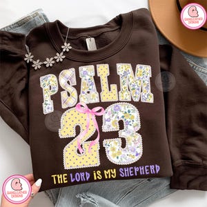 May include: A brown sweatshirt featuring the text "PSALM 23" in a floral and polka dot design. The words "THE LORD IS MY SHEPHERD" are printed below. A pink ribbon is tied around the number 2. A floral necklace is draped on the sweatshirt.