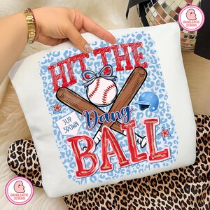 May include: White baseball-themed sweatshirt with a blue leopard print background. The design features a baseball, crossed bats, and the text "HIT THE BALL, Dang". Includes a blue baseball helmet and stars.