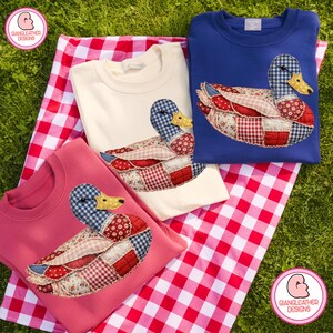 Peut inclure: Trois sweat-shirts roses, crème et bleus, chacun avec un motif de canard en patchwork. Les canards ont des têtes en vichy bleu, des ailes à motifs rouges et blancs et des becs jaunes. Les sweat-shirts sont sur une couverture de pique-nique à carreaux rouges et blancs.