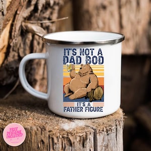 May include: White enamel camping mug with a brown bear holding a beer and sitting on a barrel. The mug has the text "It's not a dad bod, it's a father figure" in black.