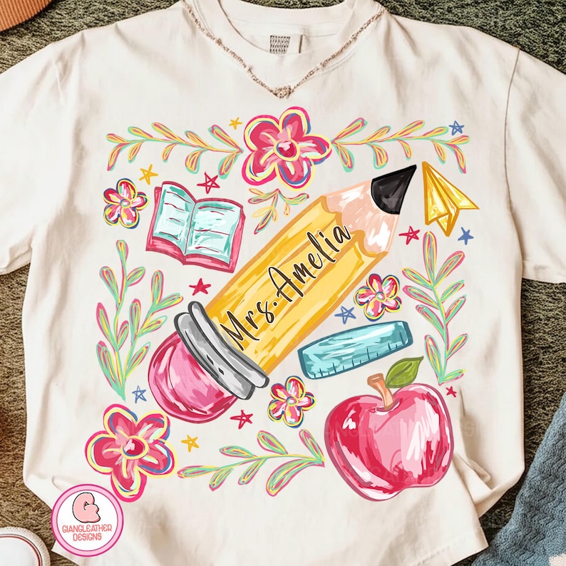 May include: Cream-colored t-shirt featuring a colorful teacher-themed design. A large yellow pencil with "Mrs. Amelia" printed on it is surrounded by flowers, a book, a ruler, a paper airplane, and a red apple. The design is whimsical and hand-drawn.