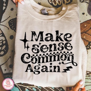 May include: A beige sweatshirt with the phrase "Make sense Common Again" in black, stylized text. The design includes stars, a checkered pattern, and lightning bolts. The sweatshirt is folded and held by hands.