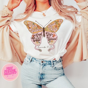 Op de afbeelding: Wit T-shirt met een roze en gele vlinderprint. De vlinder heeft de woorden "Her Children Rise Up And Call Her Blessed" en "Proverbs 31:28" erop.