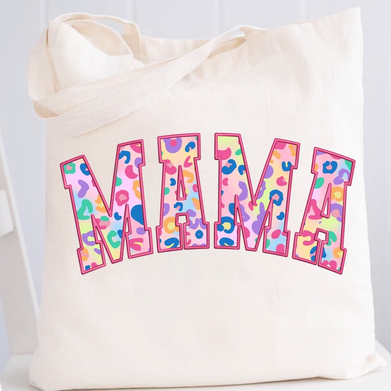 May include: A white canvas tote bag with the word "MAMA" printed in pink, outlined in black, and filled with a colorful leopard print pattern.