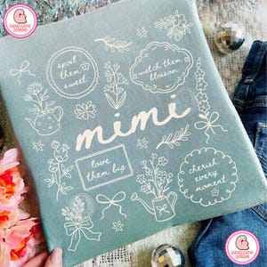 May include: A light blue fabric with white floral illustrations and the name "Mimi" in cursive. The design includes phrases like "spoil them sweet" and "cherish every moment".