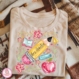 May include: Beige t-shirt with a colorful teacher-themed graphic. The design includes a yellow pencil with "Mrs. Amelia" written on it, a red apple, an open book, a ruler, and flowers. The shirt is made of soft material.