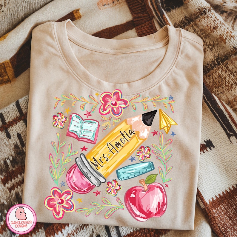 May include: Beige t-shirt with a colorful teacher-themed graphic. The design includes a yellow pencil with "Mrs. Amelia" written on it, a red apple, an open book, a ruler, and flowers. The shirt is made of soft material.