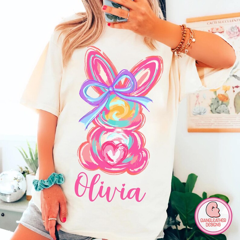 May include: A white t-shirt with a colorful abstract bunny design and the name "Olivia" printed in pink.