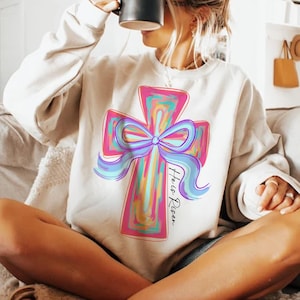 May include: A white sweatshirt with a colorful cross and bow design. The text "He is Risen" is written below the cross.