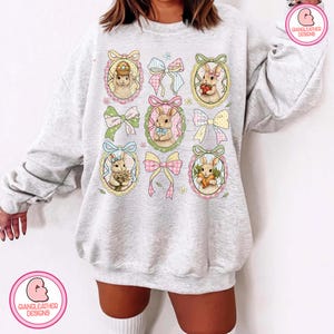 K&ouml;nnte beinhalten: Ein hellgraues Sweatshirt mit einem sich wiederholenden Muster von Osterhasen in verzierten Rahmen, durchsetzt mit bunten Schleifen. Die Hasen halten Ostereier, Blumen und Karotten. Das Design ist in Pastellfarben gehalten und erzeugt einen skurrilen und festlichen Look.
