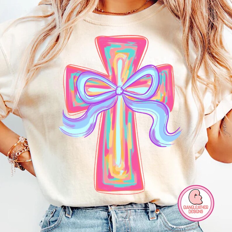 May include: A white t-shirt with a colorful cross design featuring a large purple bow. The cross is painted with pink, blue, yellow, and orange colors. The text "Giangleather Designs" is visible in the bottom right corner.
