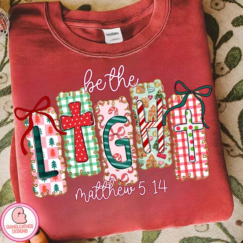 May include: A maroon sweatshirt featuring the phrase "be the LIGHT" in a decorative font. The letters are designed with Christmas-themed patterns, including crosses, candy canes, and gingerbread houses. The text "matthew 5:14" is below.