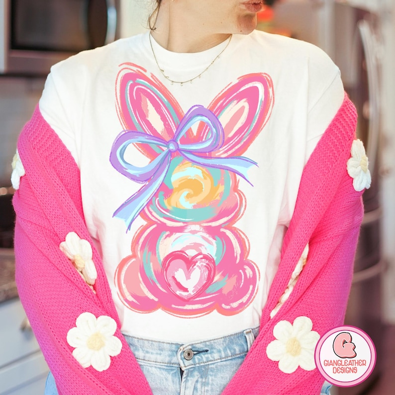 May include: A white t-shirt with a colorful abstract bunny design. The bunny has a large blue bow and a pink heart on its chest.