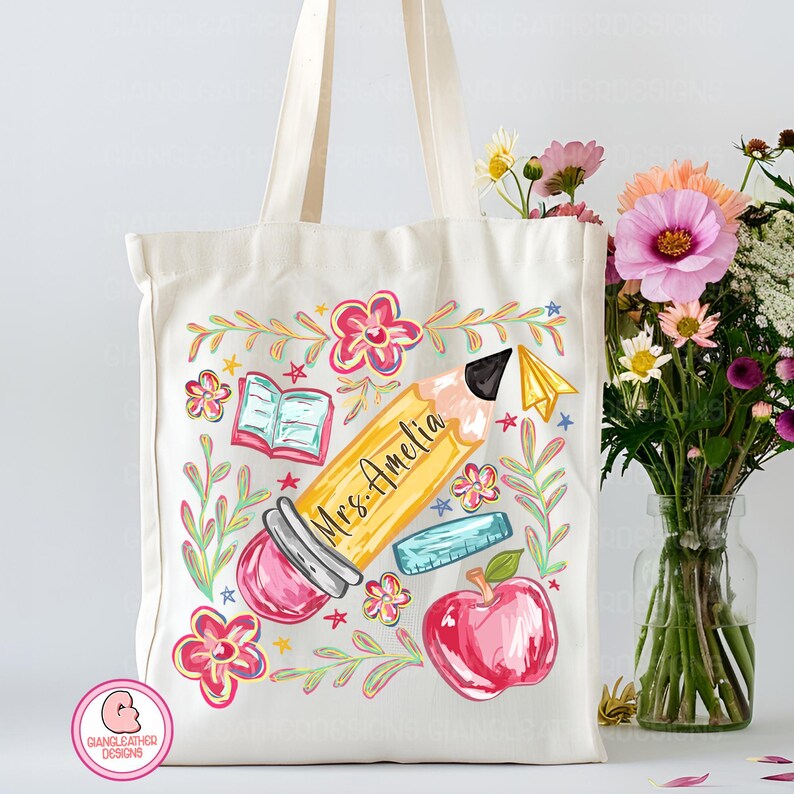May include: Cream-colored tote bag featuring a colorful school-themed design. A large yellow pencil with "Mrs. Amelia" written on it is the focal point, surrounded by flowers, an apple, an eraser, and an open book. The bag has cream-colored handles.