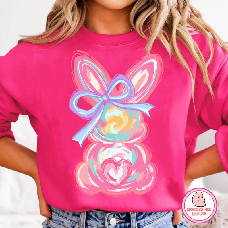 May include: A pink sweatshirt with a colorful abstract bunny design. The bunny has a blue bow and a pink heart.