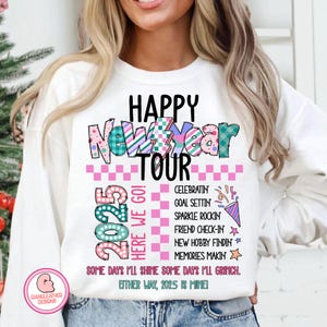 May include: White sweatshirt with a colorful graphic design that says "Happy New Year Tour 2025" with a pink and white checkered background. The graphic also includes the text "Here We Go!" and a list of activities for the new year, including "Celebratin, Goal Settin, Sparkle Rockin, Friend Check-In, New Hobby Findin, Memories Makin". The graphic ends with the text "Some days I'll shine some days I'll Grinch. Either way, 2025 is mine!"