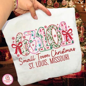 May include: White t-shirt with a festive Christmas design. The design features the numbers "63101" in a decorative font, filled with patterns of Christmas trees, holly, and snowflakes. The text "Small Town Christmas ST. LOUIS, MISSOURI" is printed below.