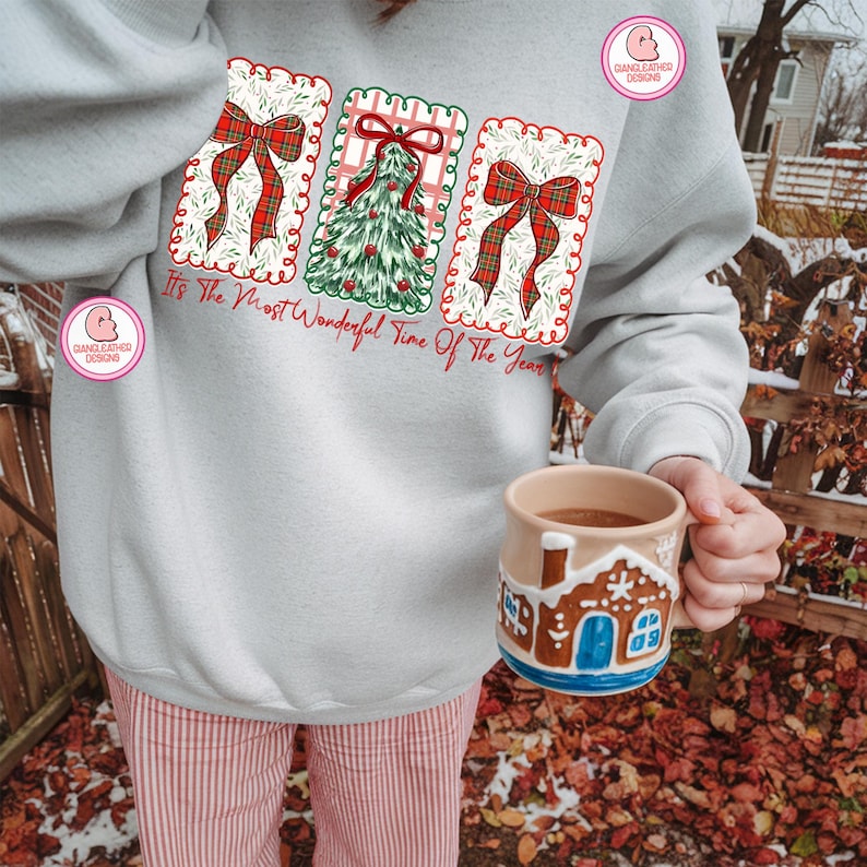 May include: A light gray sweatshirt with a Christmas-themed design featuring red bows and a Christmas tree. The text "It's The Most Wonderful Time Of The Year" is printed below the design. A gingerbread house mug is held in the hand.