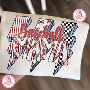 May include: White t-shirt with the words "Baseball MAMA" in red script. The letters are filled with baseball stitching and are set against a lightning bolt design with red and white stripes, blue accents, and a black and white checkered pattern.