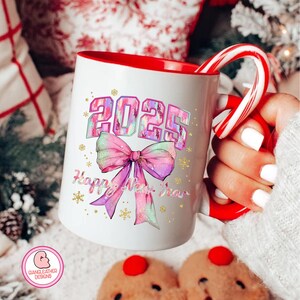 May include: White ceramic mug with a red rim and handle. The mug features a colorful design with the year 2025 in a glittery font and a pink bow with a glittery design. The text "Happy New Year" is written below the bow.