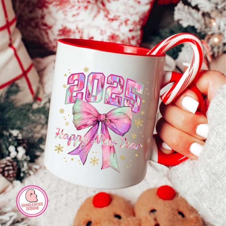 May include: White ceramic mug with a red rim and handle. The mug features a colorful design with the year 2025 in a glittery font and a pink bow with a glittery design. The text "Happy New Year" is written below the bow.