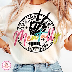 May include: Cream-colored t-shirt with a graphic that reads "PLEASE HOLD I'M MENTALLY BUFFERING." The design features a black skeleton hand making a peace sign, a loading bar, and colorful text.