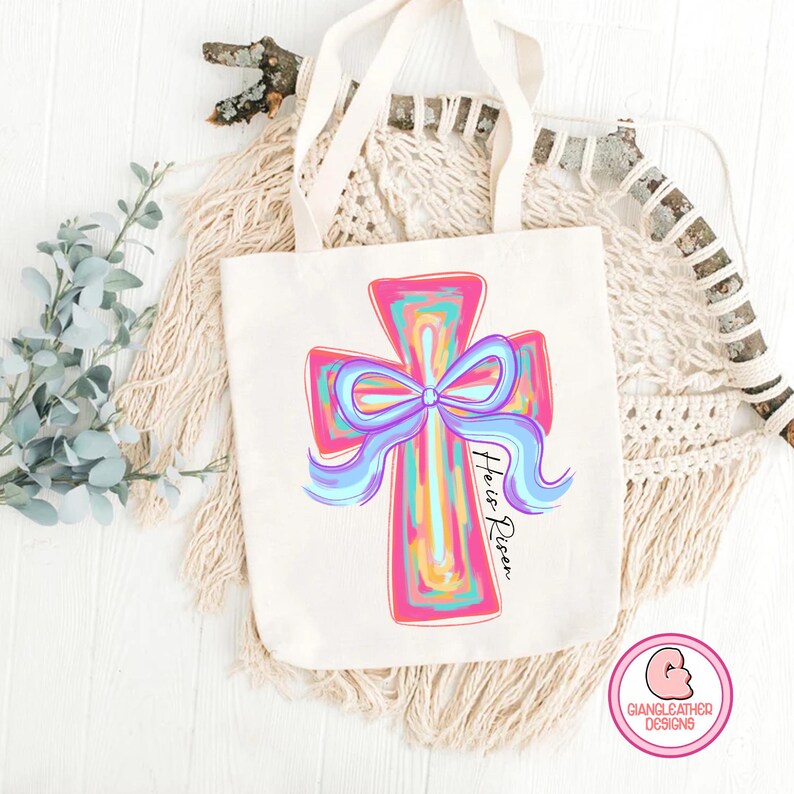 May include: A white canvas tote bag with a pink and blue cross with a blue bow and the text "He is Risen".