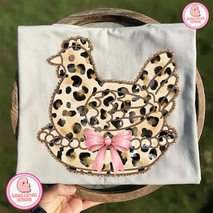 May include: Cream-colored t-shirt featuring a hen design with a leopard print pattern. The hen is adorned with a pink bow and sits on a nest. The design is outlined in brown.