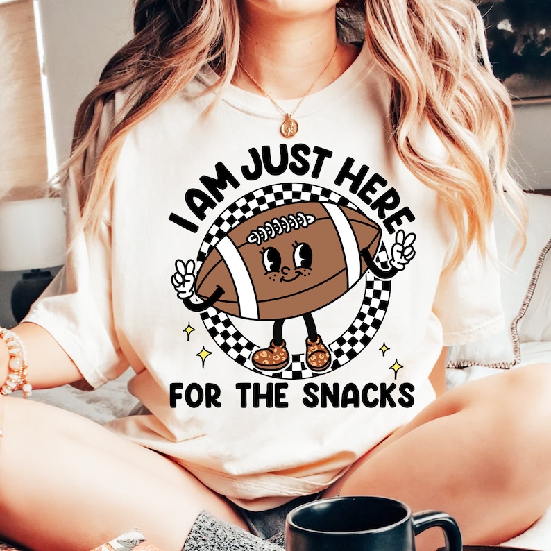May include: White t-shirt with a cartoon football character wearing shoes and holding peace signs. The text on the shirt reads "I am just here for the snacks".